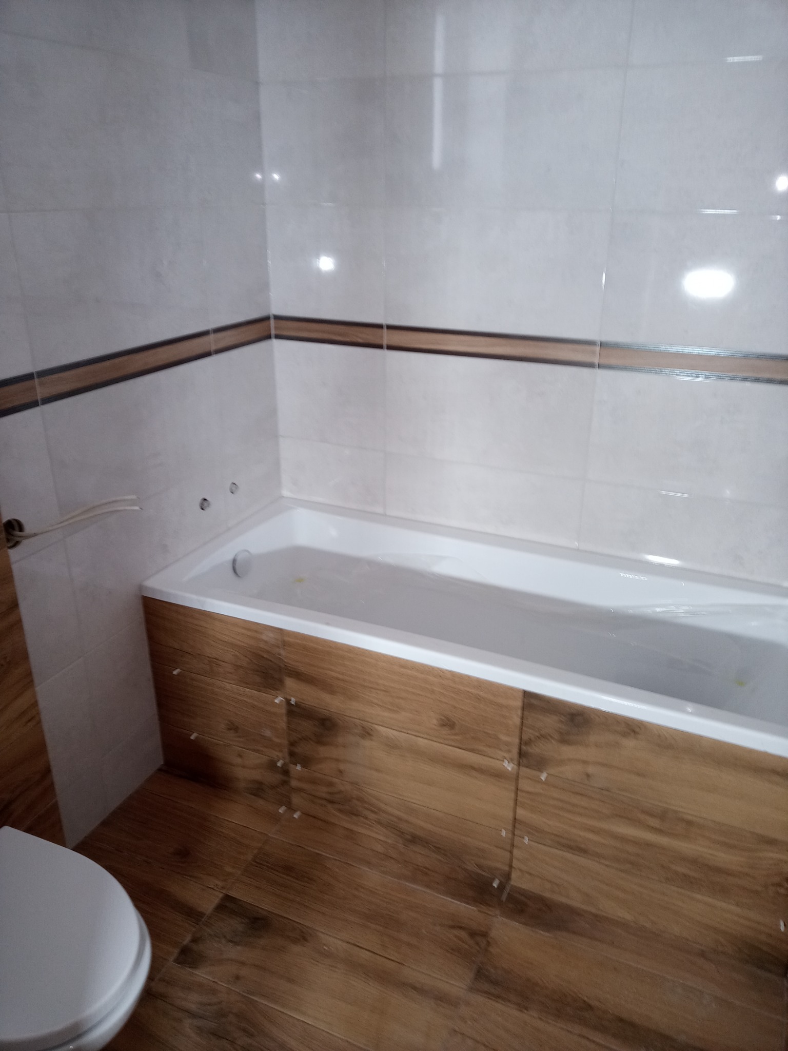 Wykończona łazienka z biało-drewnianymi płytkami, obudowaną wanną i częściowo widoczną toaletą, widoczne plastikowe krzyżyki dystansowe między płytkami.