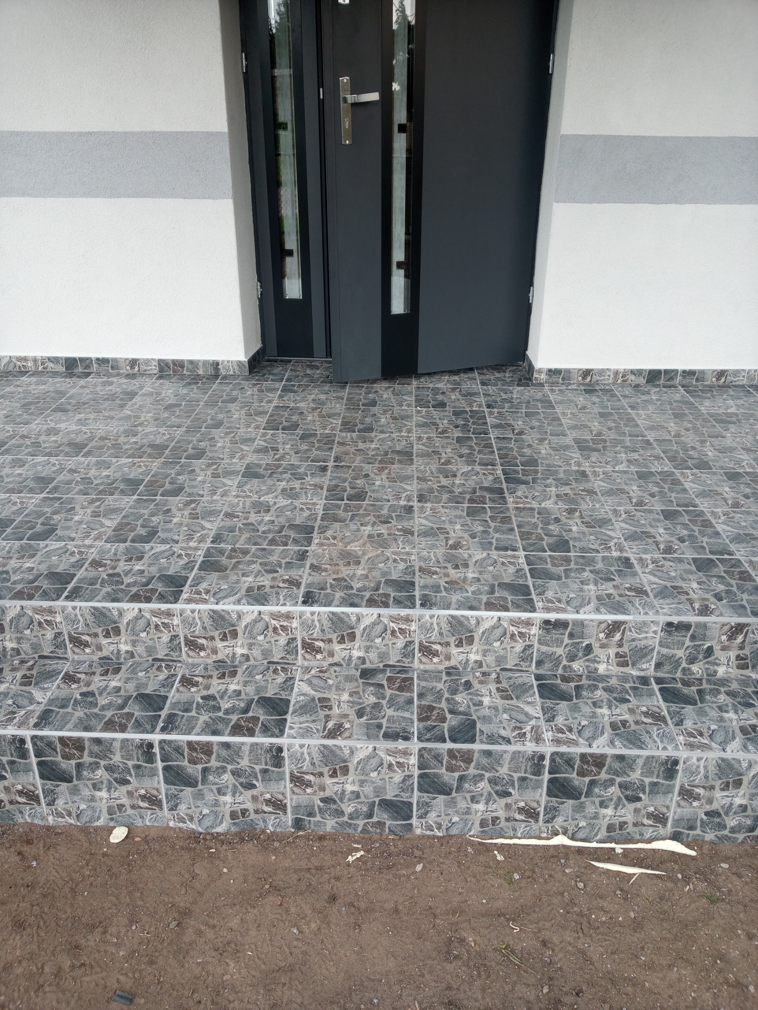 Wykończony taras przed wejściem do domu z szarymi płytkami imitującymi kamień, widoczne dwa stopnie i częściowo otwarte, grafitowe drzwi wejściowe.