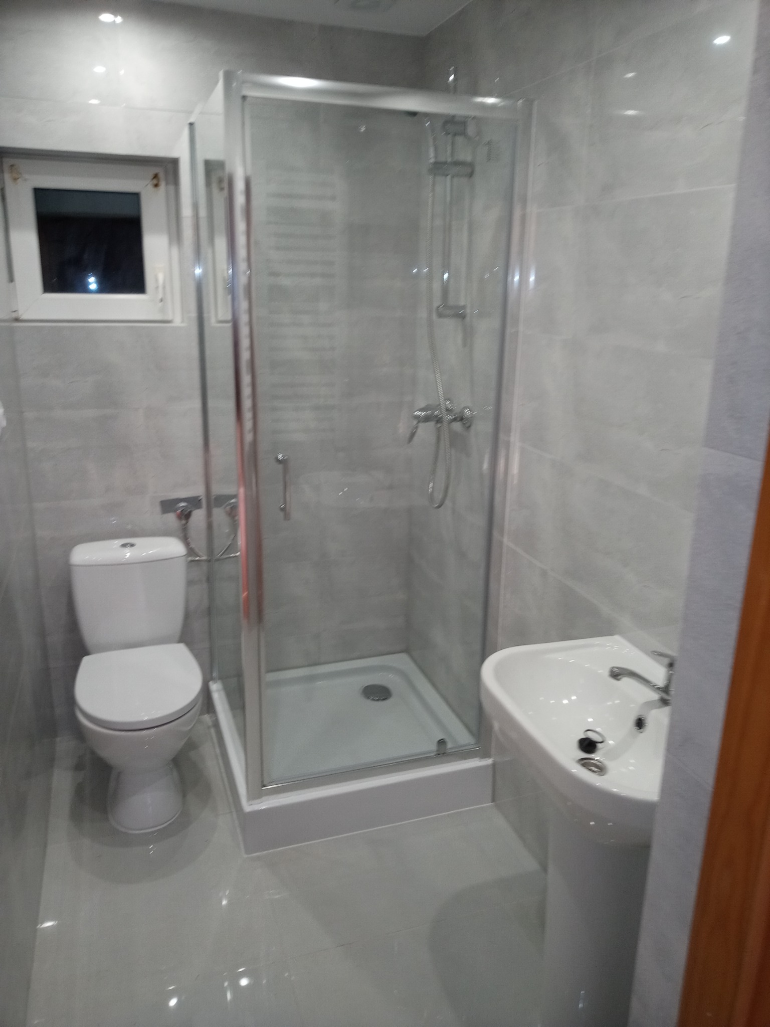 Nowoczesna łazienka z szarymi płytkami, kabiną prysznicową z przezroczystego szkła, białą umywalką na postumencie i toaletą. Widoczne okno z białymi ramami.