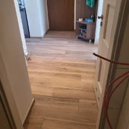 Ułożona podłoga z płytek imitujących drewno w korytarzu, widoczne przewody elektryczne przy futrynie drzwi.