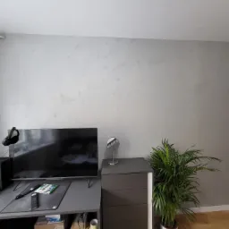 Ściana w pokoju po szpachlowaniu z efektem dekoracyjnym, widoczne rury grzewcze pod sufitem, biurko z elektroniką i roślina doniczkowa.