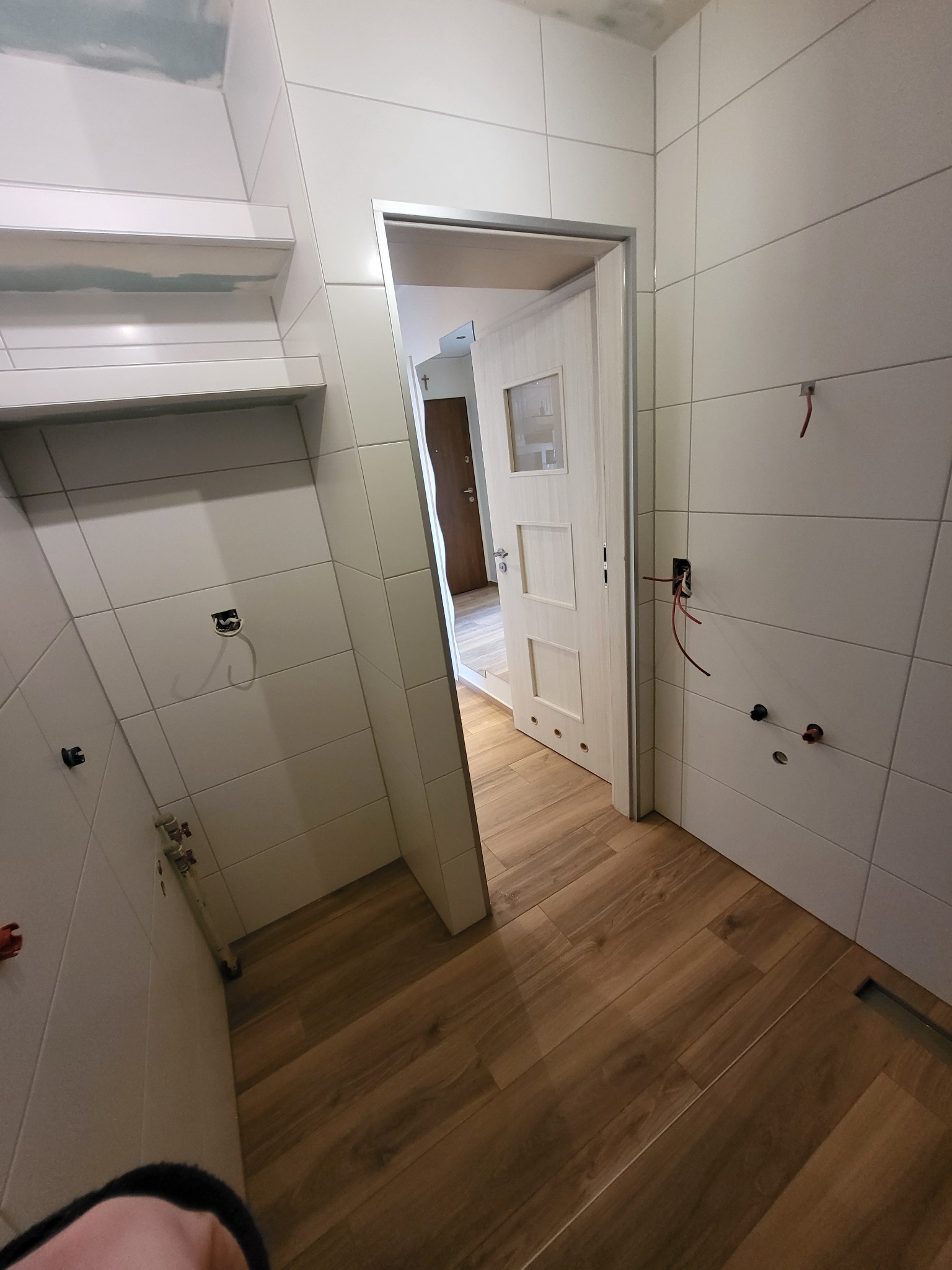 Wnętrze łazienki w trakcie remontu, widoczne białe płytki na ścianach, drewnopodobne panele na podłodze, niezakończone prace instalacyjne i fragment otwartych drzwi do innego pomieszczenia.