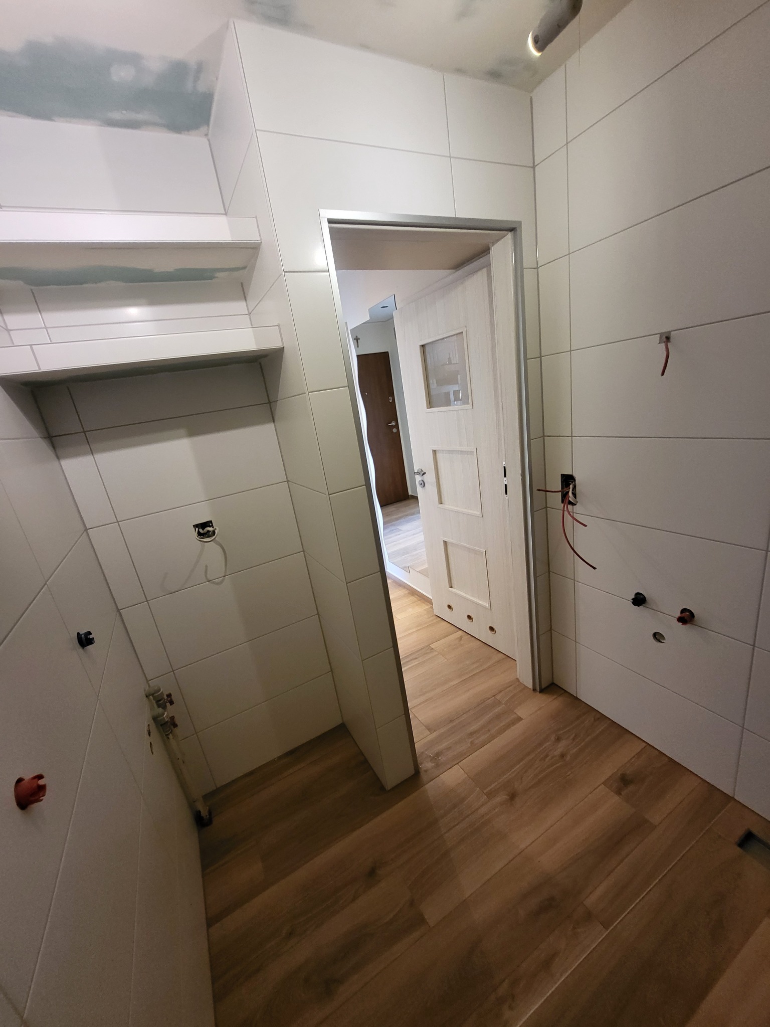 Wnętrze łazienki w trakcie prac wykończeniowych: białe płytki ścienne, drewnopodobne panele podłogowe, widoczne kable i puszki elektryczne, niezakończone szpachlowanie sufitu, otwarte drzwi...