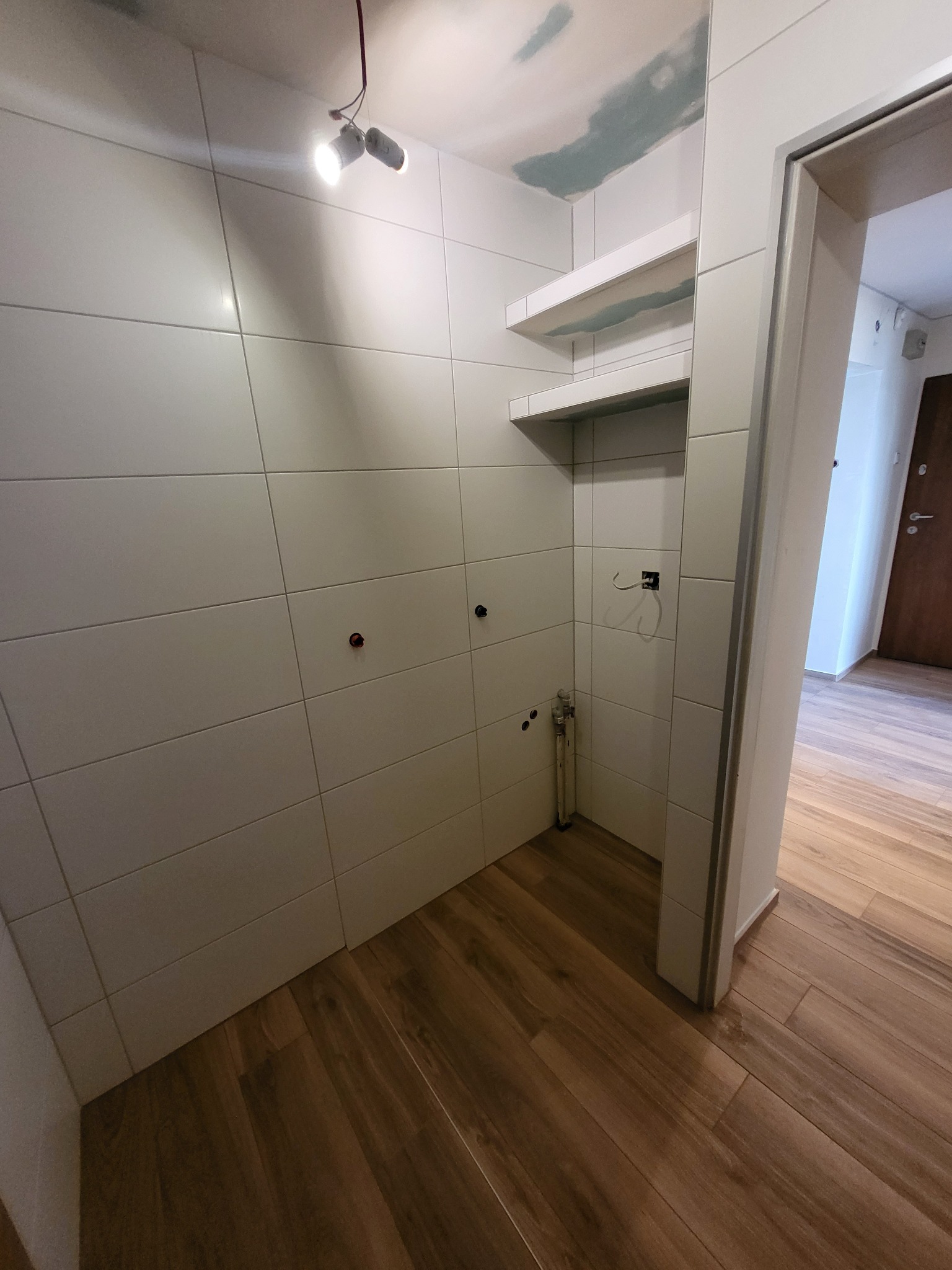 Wnętrze łazienki w trakcie prac wykończeniowych: białe, poziome płytki na ścianach, przygotowane punkty instalacyjne, drewnopodobna podłoga, widoczne półki i fragment sufitu z niedokończonymi pracami.