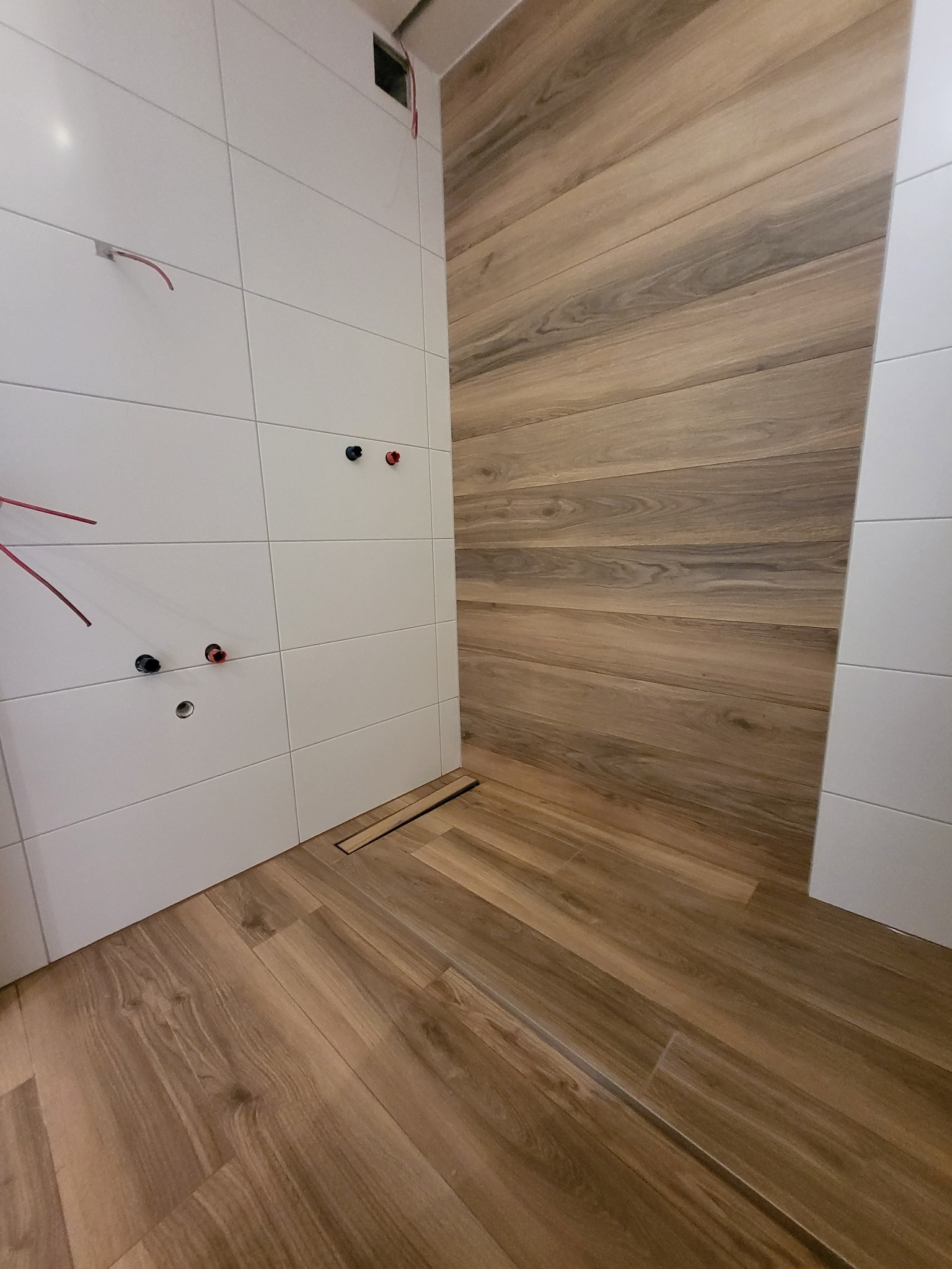 Wnętrze łazienki w trakcie wykańczania, z białymi płytkami na ścianie z instalacją hydrauliczną i elektryczną oraz płytkami imitującymi drewno na podłodze i fragmencie ściany, widoczny odpływ liniowy.