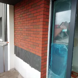 Fragment ściany budynku z nową elewacją z czerwonej cegły klinkierowej, widoczny fragment ściany z nałożonym klejem oraz okno zabezpieczone folią ochronną.