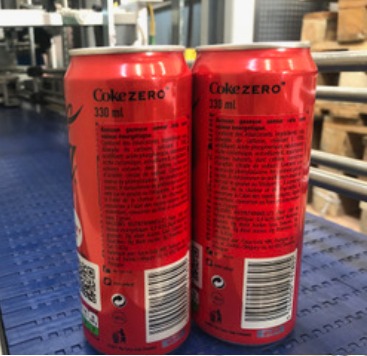 Dwie czerwone puszki Coca-Cola Zero 330 ml ustawione obok siebie na linii produkcyjnej, widoczne etykiety z drobnym drukiem i kodami kreskowymi, tło stanowią elementy linii produkcyjnej.