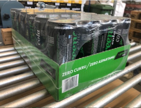 Zafoliowany karton z puszkami napoju gazowanego na metalowym przenośniku rolkowym, widoczny napis 'ZERO CUKRU / ZERO ASPARTAMU'.