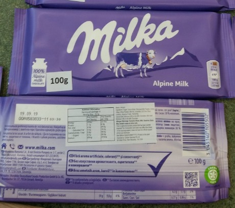 Stosy fioletowych opakowań czekolady Milka Alpine Milk 100g, widoczne logo z krową na tle górskim.