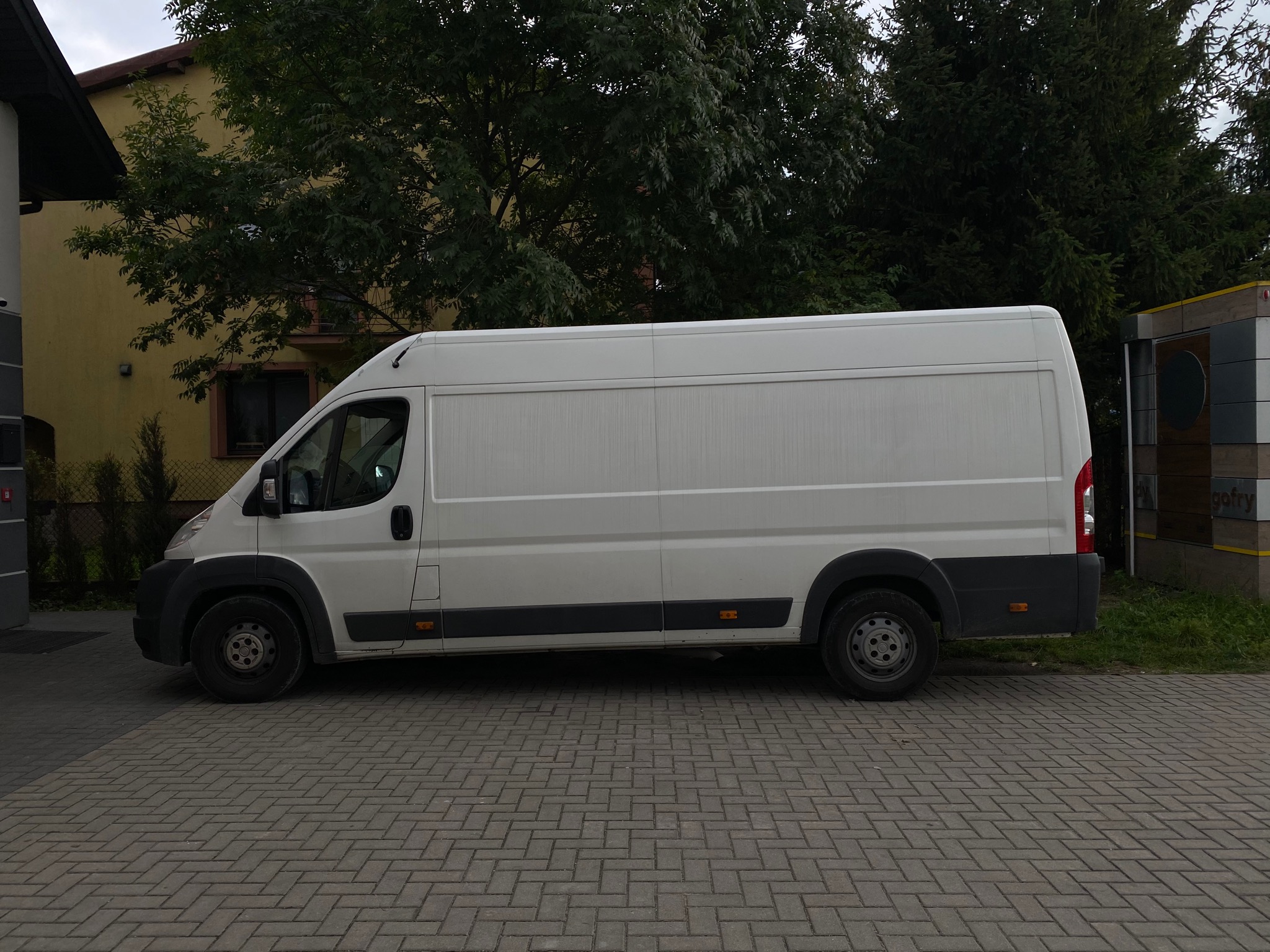 Biały furgon dostawczy zaparkowany na brukowanej drodze przed budynkiem i drzewami.