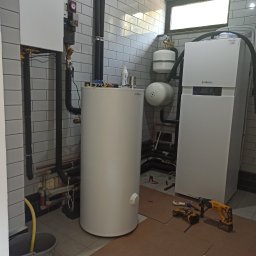 Santehservis - Instalacja urządzeń grzewczych Viessmann w kotłowni wyłożonej białymi płytkami, widoczne pompy ciepła, zbiornik buforowy, instalacja rurowa, narzędzia pracy i kartonowe zabezpieczenie podłogi.