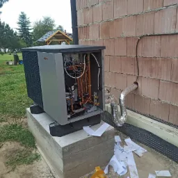 Zewnętrzna jednostka pompy ciepła Viessmann podczas instalacji przy ścianie budynku z bloczk&oacute;w, widoczne miedziane rury i przewody, fragment elewacji i trawnika, porozrzucane fragmenty izolacji.