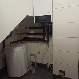Santehservis - Kocioł grzewczy na ścianie z podłączonymi rurami, obok biały zasobnik na wodę zabezpieczony folią, widoczne płytki na ścianach i podłodze, instalacja w trakcie montażu.