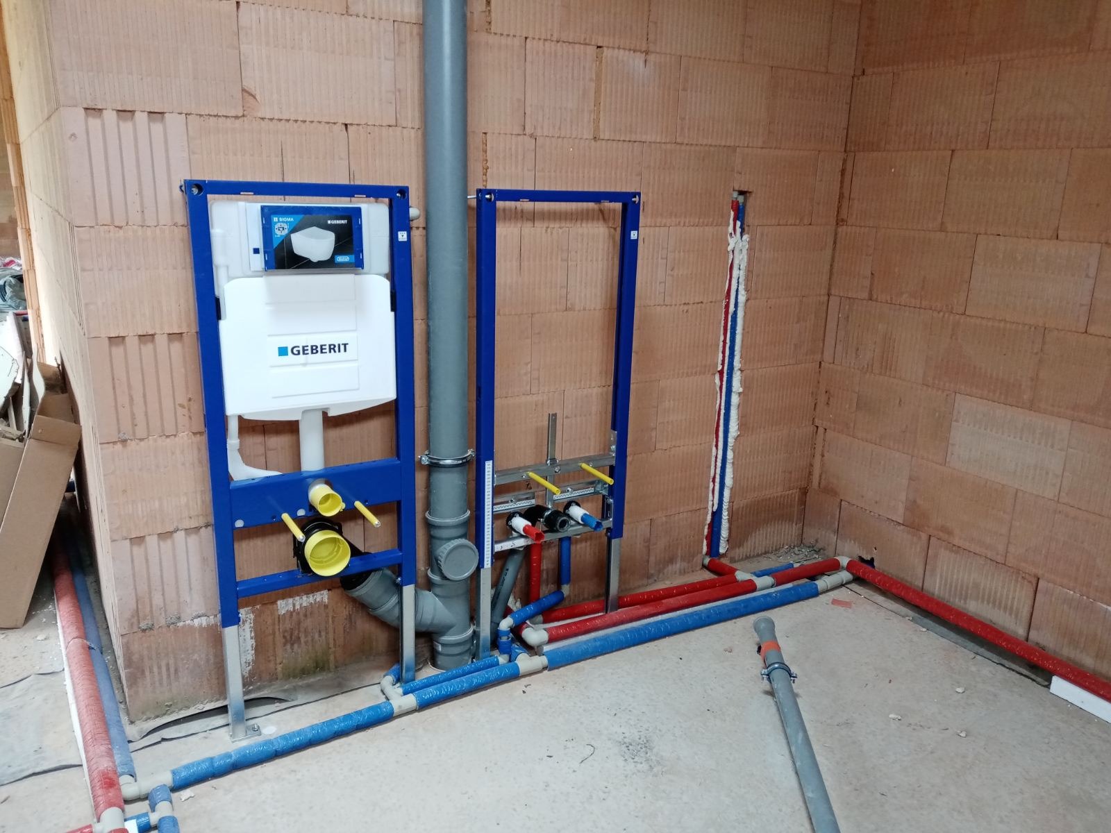 Montaż stelaży podtynkowych Geberit z instalacją wodną w surowym wnętrzu z ceglanymi ścianami i betonową podłogą, widoczne rury w izolacji oraz elementy mocujące.