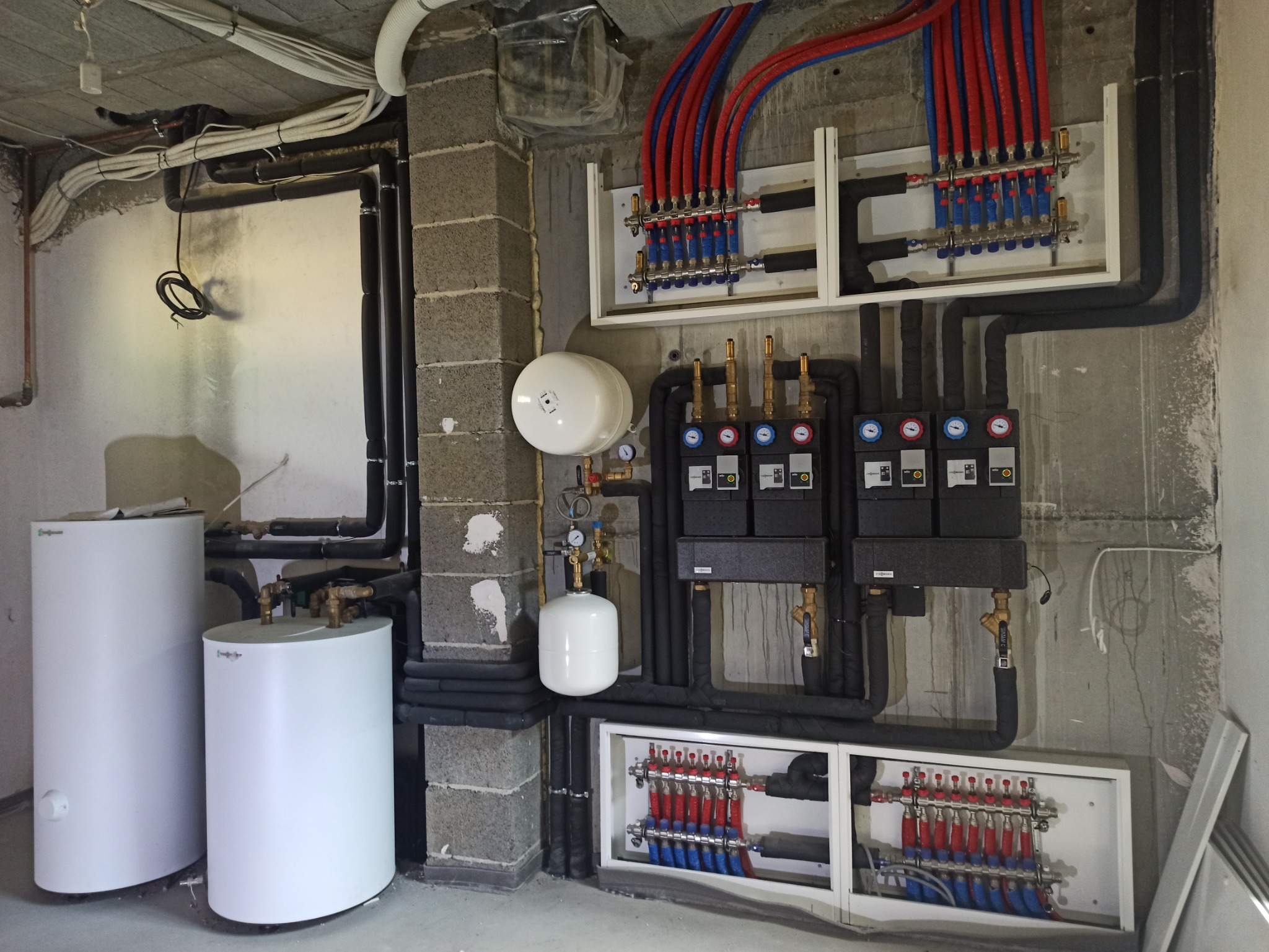 Rozdzielacze ogrzewania podłogowego z rurami w kolorze czerwonym i niebieskim, zbiorniki wyrównawcze ciśnienia oraz automatyka kotłowni w surowym pomieszczeniu z betonowymi ścianami.