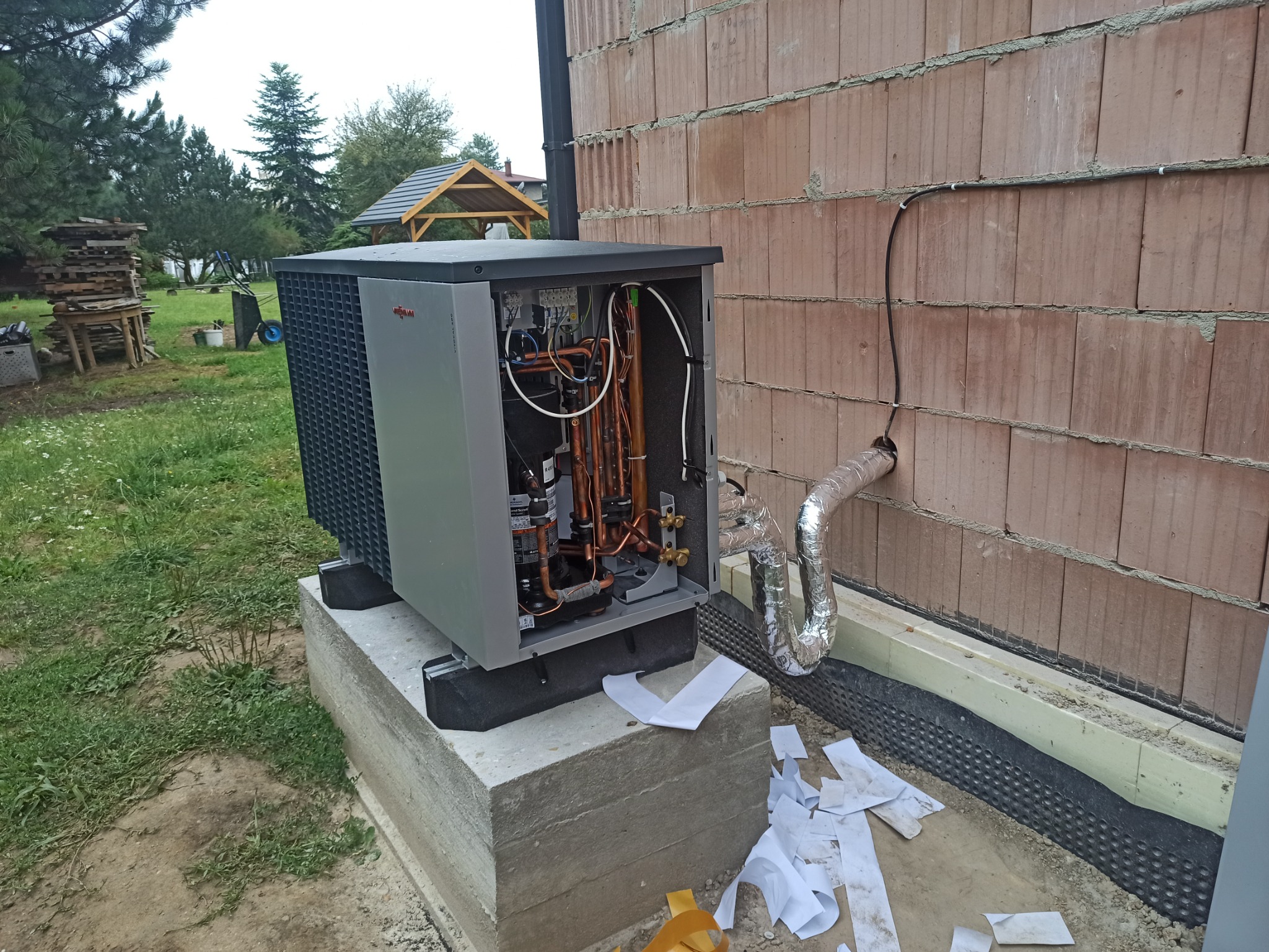 Zewnętrzna jednostka pompy ciepła Viessmann podczas instalacji przy ścianie budynku z bloczków, widoczne miedziane rury i przewody, fragment elewacji i trawnika, porozrzucane fragmenty izolacji.