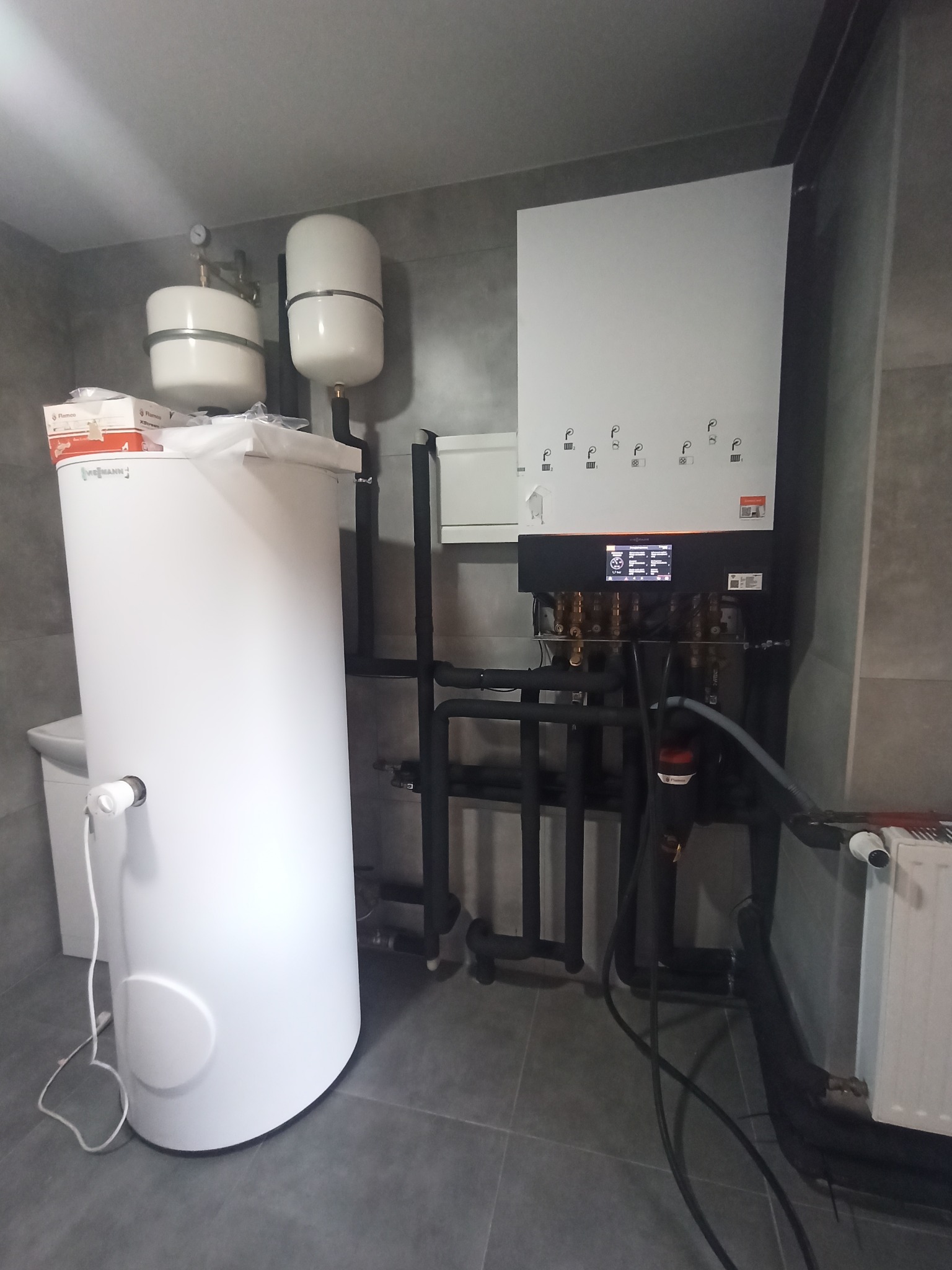 Nowoczesna kotłownia z białym zasobnikiem ciepłej wody, czarnymi rurami instalacyjnymi i szarą glazurą na ścianach, widoczne elementy automatyki i manometry.