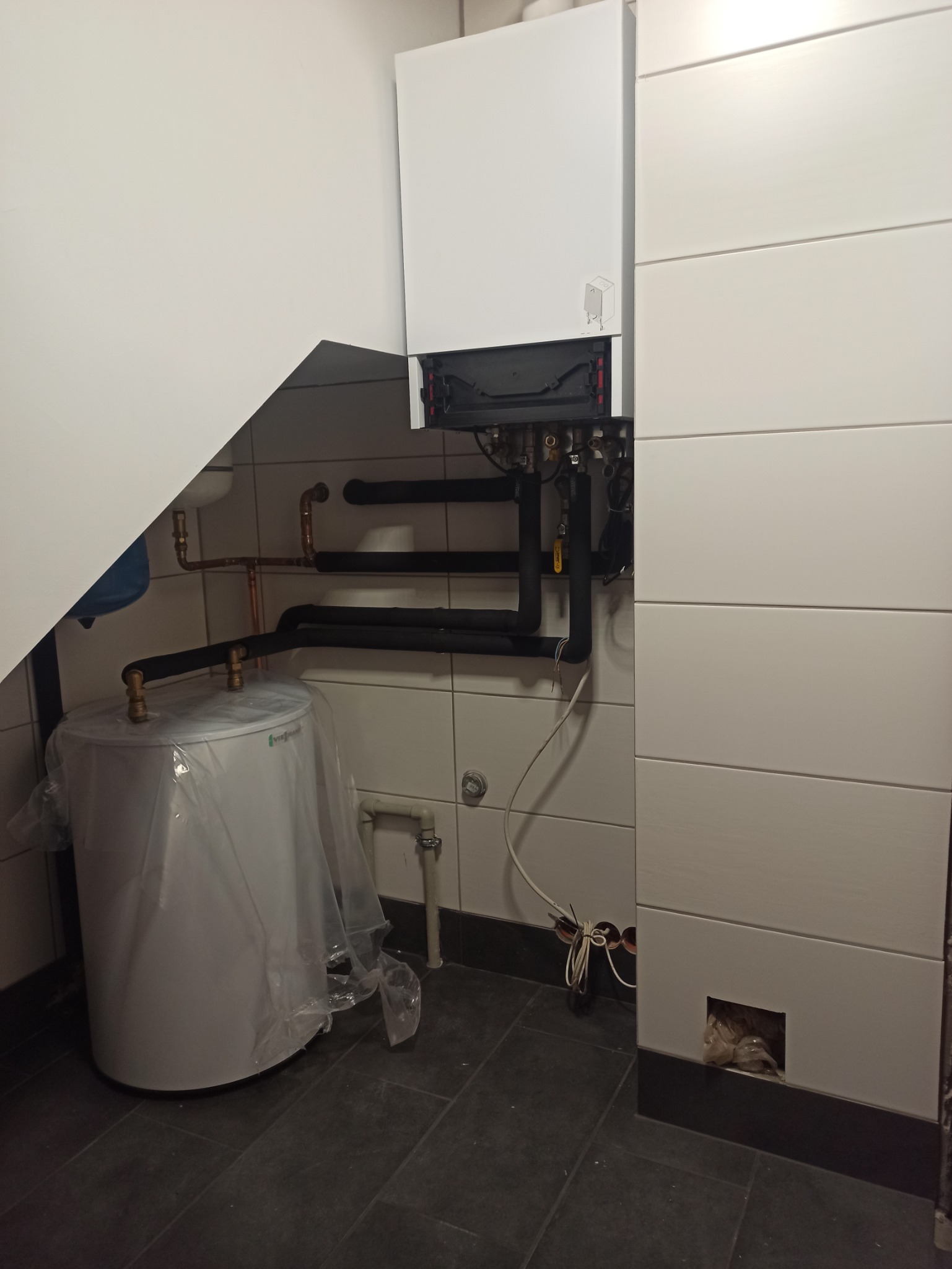 Kocioł grzewczy na ścianie z podłączonymi rurami, obok biały zasobnik na wodę zabezpieczony folią, widoczne płytki na ścianach i podłodze, instalacja w trakcie montażu.