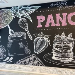 ELERTs DESIGN - Tablica kredowa z rysunkami białych konturów: ananas, ubijaczka, worek z ziarnami, cytryna, gofr, truskawka i stos naleśników z owocami, napis 'PANCA' w kolorze różowym, głośnik Skytec w lewym...