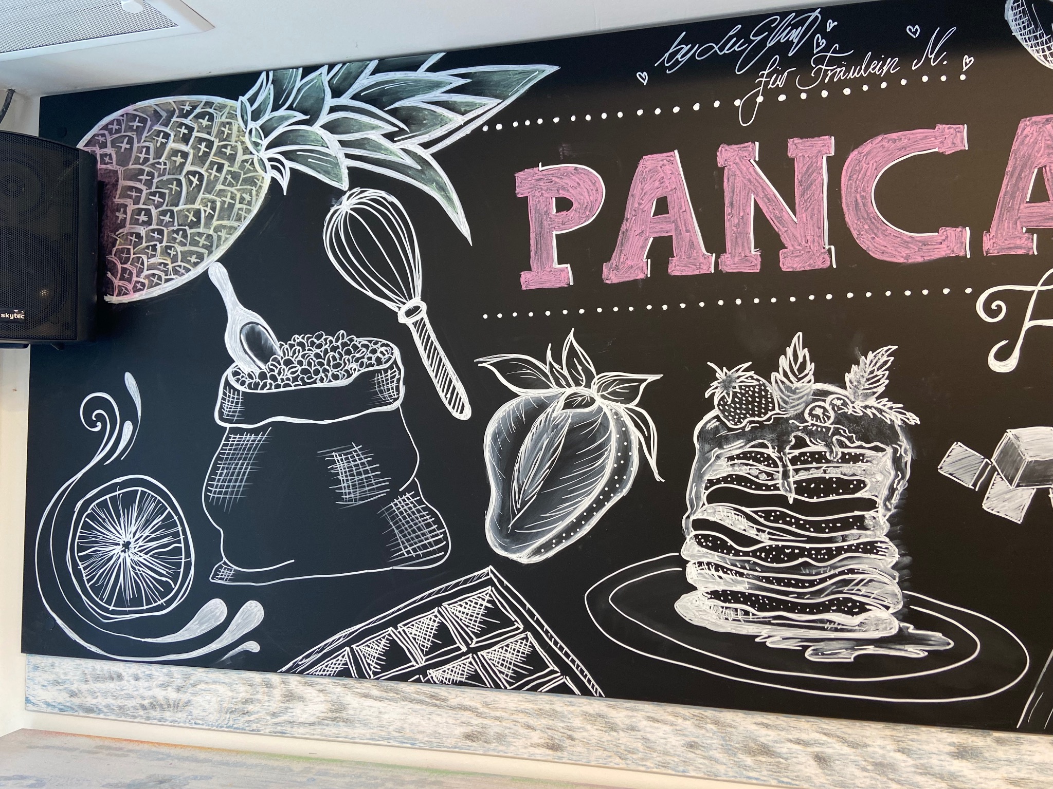 Tablica kredowa z rysunkami białych konturów: ananas, ubijaczka, worek z ziarnami, cytryna, gofr, truskawka i stos naleśników z owocami, napis 'PANCA' w kolorze różowym, głośnik Skytec w lewym...