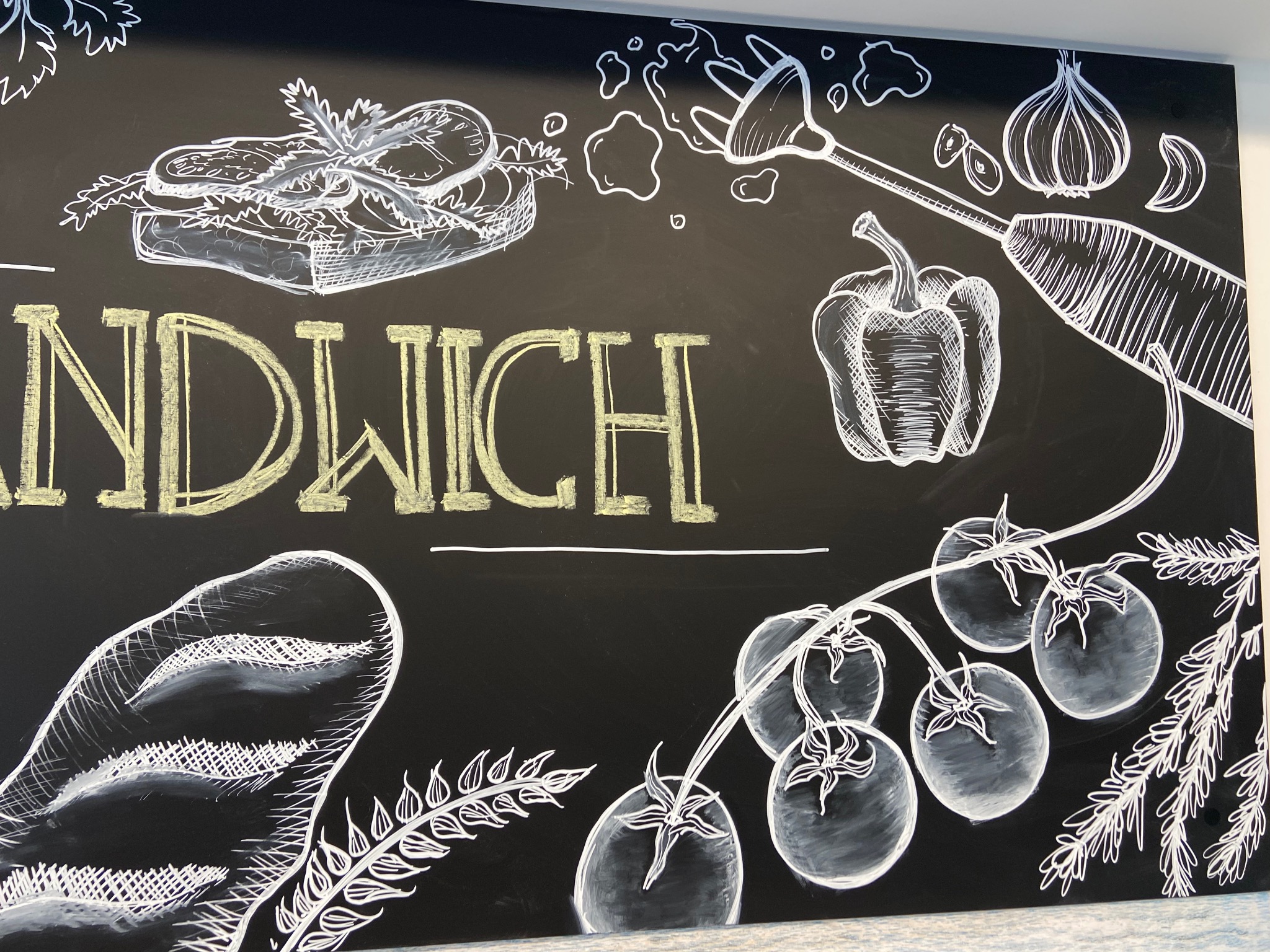 Tablica kredowa z rysunkiem kanapki, warzyw i napisem 'SANDWICH' wykonanym żółtą kredą, stylizowana na menu restauracji.