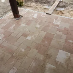 Ułożona kostka brukowa w odcieniach beżu i czerwieni, widoczny fragment drewnianej konstrukcji i piasek na powierzchni kostki.