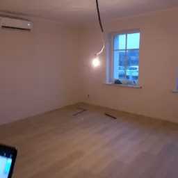 Puste pomieszczenie z nową podłogą panelową, widoczne dwa okna z zewnętrznym widokiem, klimatyzator na ścianie, wisząca żarówka, telefon w dolnym rogu kadru.