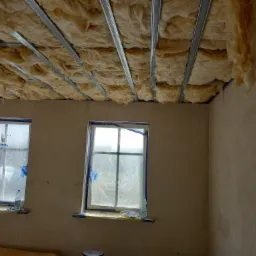 Wnętrze pokoju w trakcie prac ociepleniowych: wełna mineralna zamontowana między metalowymi profilami sufitowymi, okna zabezpieczone folią malarską, widoczne ślady po szpachlowaniu na ścianach.
