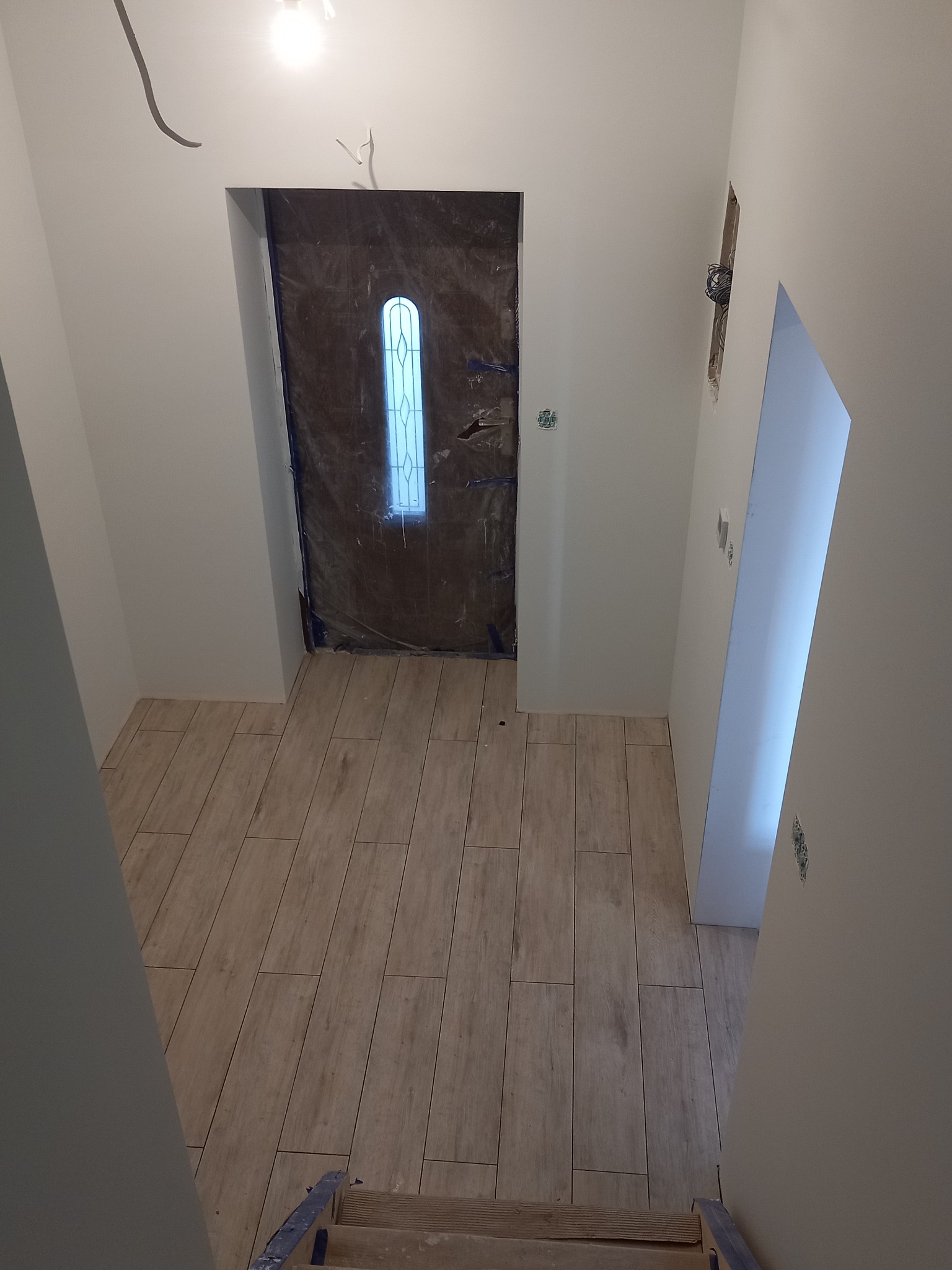 Wnętrze w trakcie remontu: świeżo pomalowane białe ściany, nowa podłoga z paneli imitujących drewno, zabezpieczone folią drzwi wejściowe z szybą, widoczne niedokończone instalacje elektryczne.
