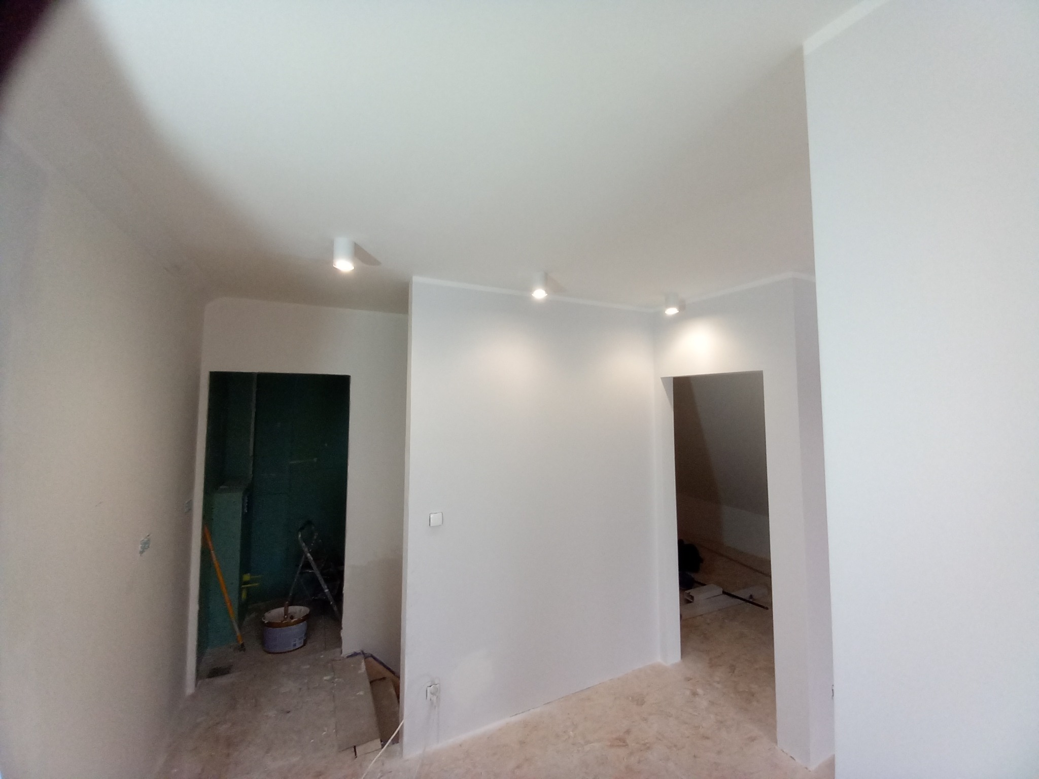 Wnętrze po malowaniu ścian na biało, widoczne trzy reflektory sufitowe, drzwi do pomieszczenia z drabiną i wiaderkiem, ślady prac remontowych na podłodze.