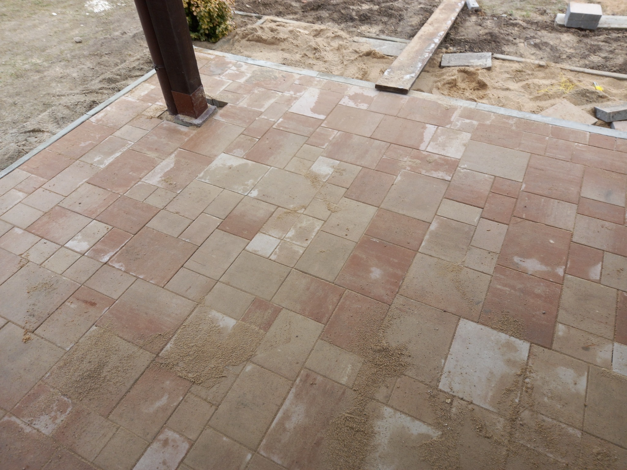 Ułożona kostka brukowa w odcieniach beżu i czerwieni, widoczny fragment drewnianej konstrukcji i piasek na powierzchni kostki.