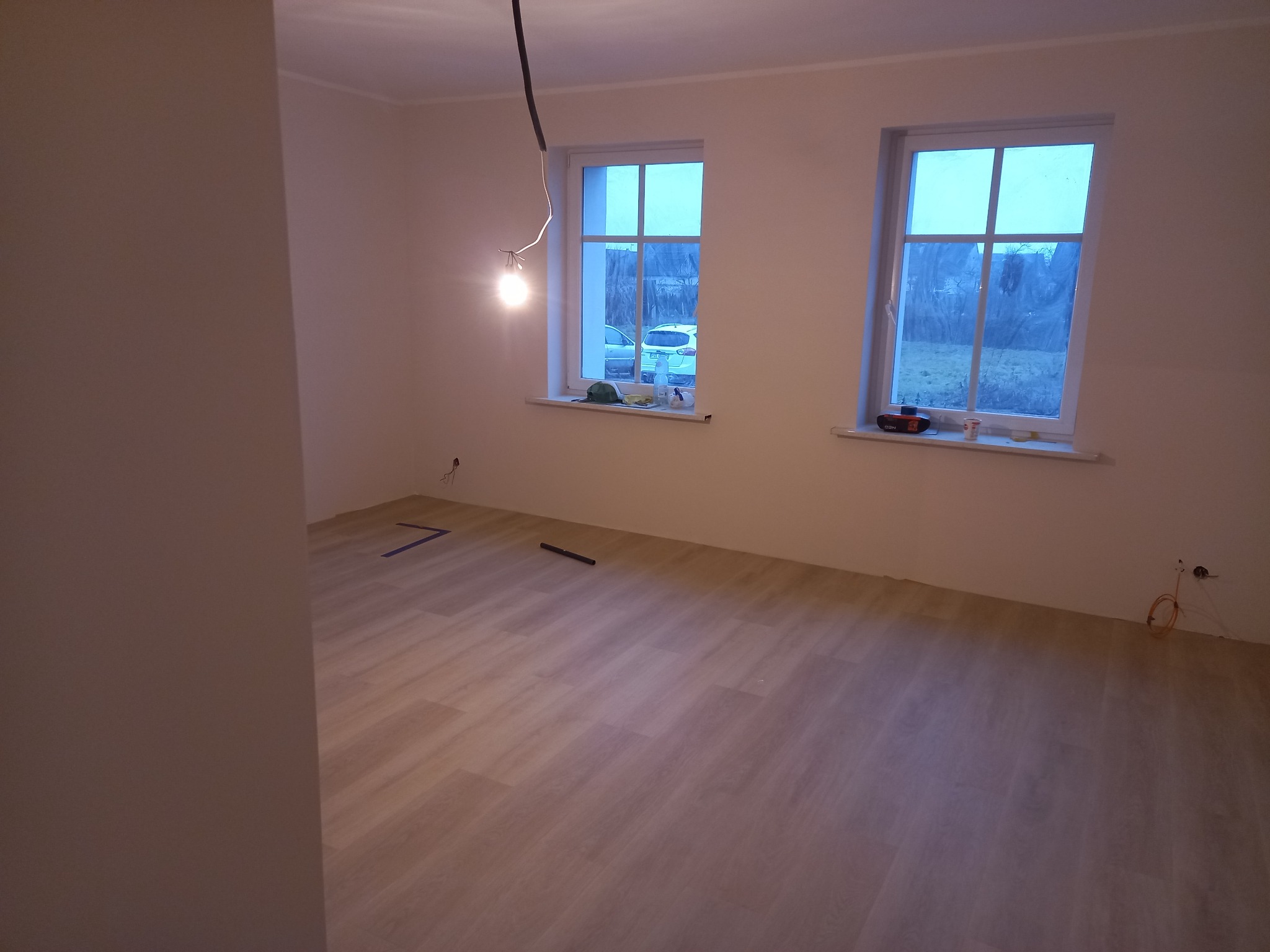 Pokój po remoncie z nową, jasną podłogą z paneli, widoczne dwa okna z białymi ramami, surowa żarówka zwisająca z sufitu, ślady taśmy malarskiej na podłodze.