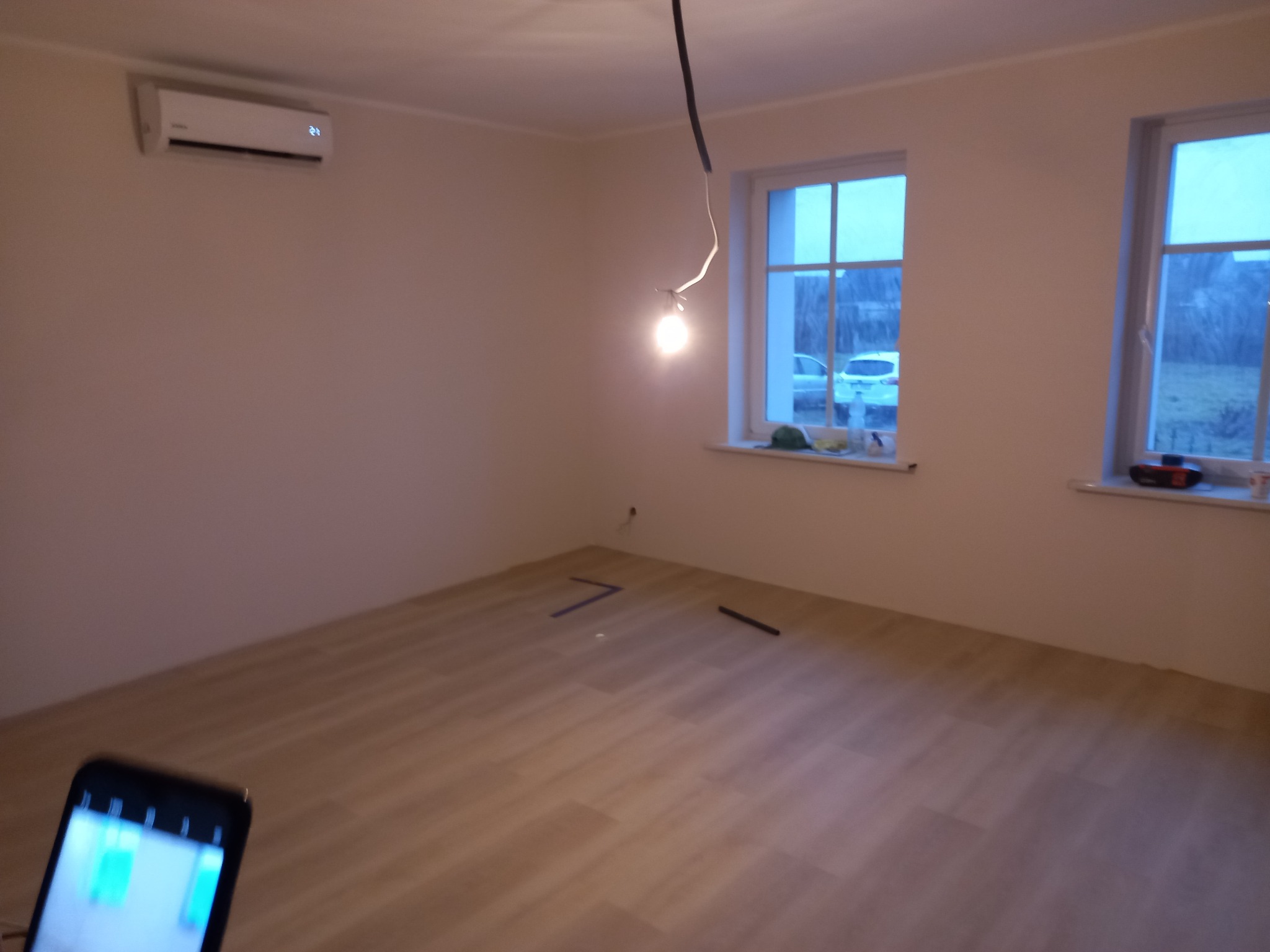 Puste pomieszczenie z nową podłogą panelową, widoczne dwa okna z zewnętrznym widokiem, klimatyzator na ścianie, wisząca żarówka, telefon w dolnym rogu kadru.