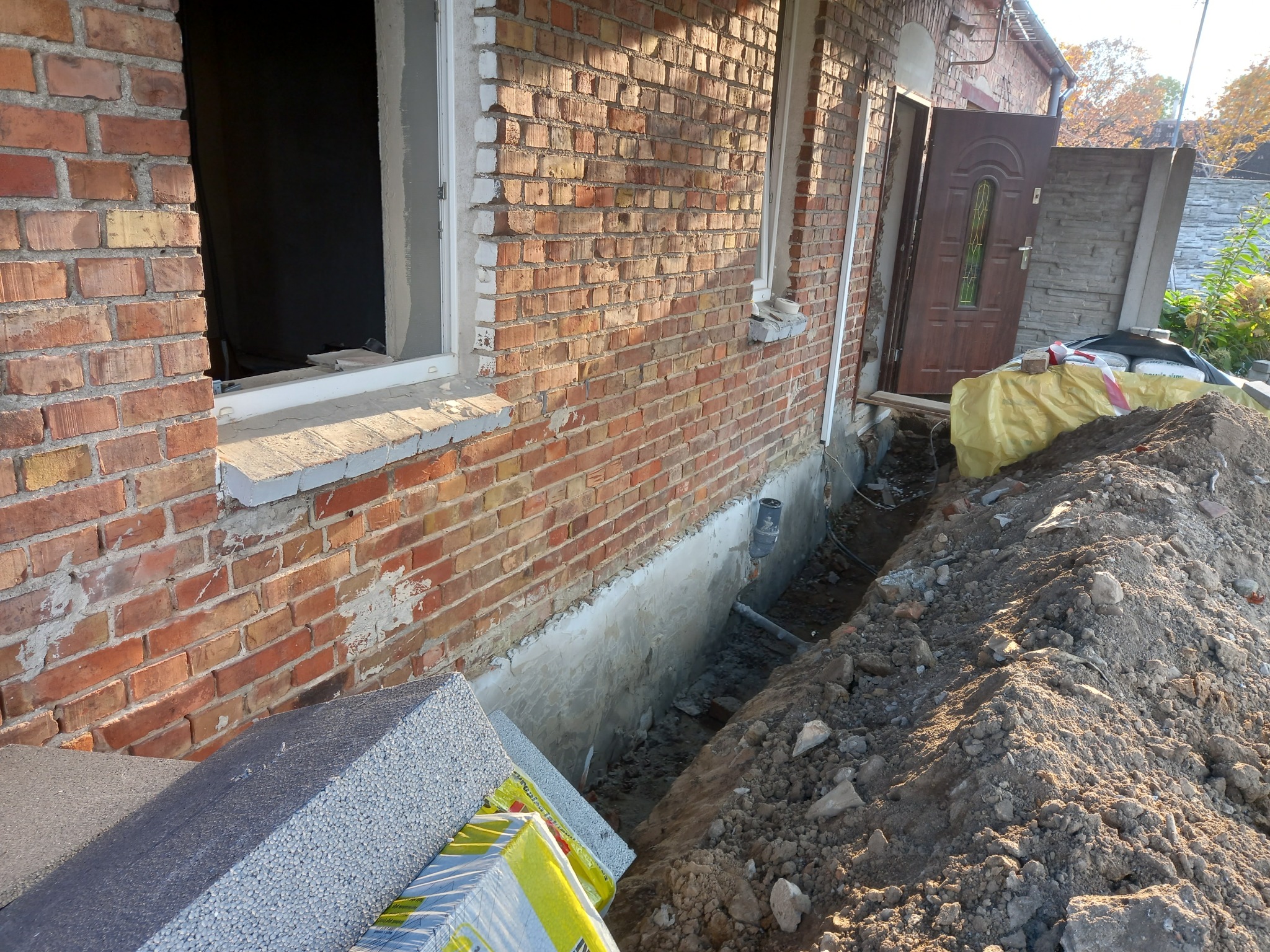 Ceglana ściana budynku z odsłoniętym fundamentem, przygotowanym do ocieplenia, widoczne materiały izolacyjne i wykop