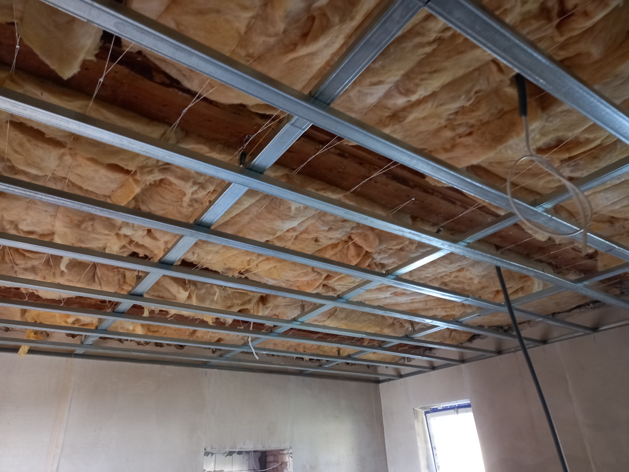 Sufit podwieszany w trakcie montażu: metalowa konstrukcja rusztu, ocieplenie wełną mineralną, widoczne przewody i fragment ściany z oknem.
