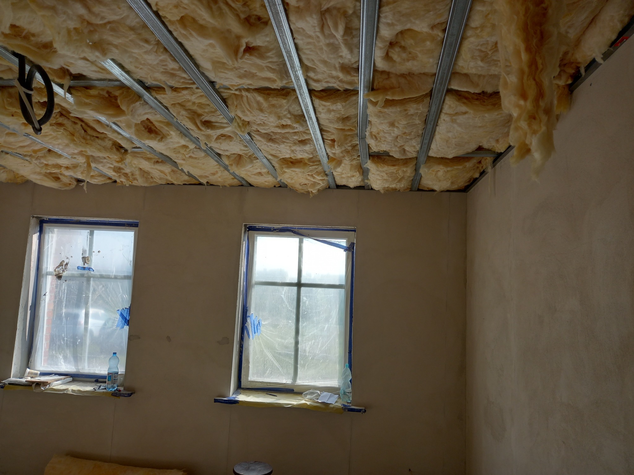 Wnętrze pokoju w trakcie prac ociepleniowych: wełna mineralna zamontowana między metalowymi profilami sufitowymi, okna zabezpieczone folią malarską, widoczne ślady po szpachlowaniu na ścianach.