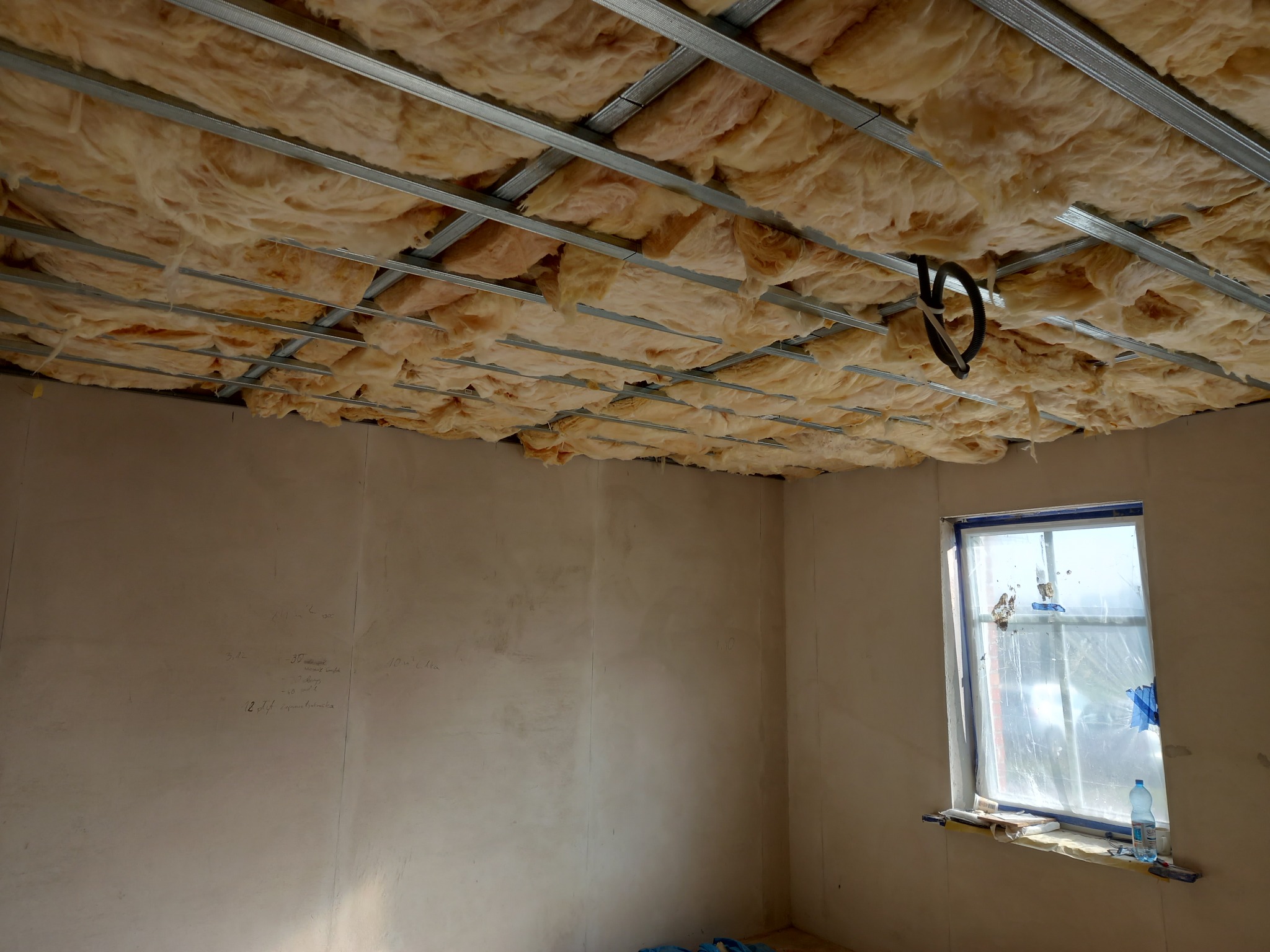 Izolacja z wełny mineralnej zamontowana w metalowym stelażu sufitu podwieszanego, widok na okno z folią ochronną i butelką wody na parapecie.