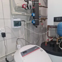 Węzeł cieplny z białym zmiękczaczem wody Viessmann Aquahome Duo Smart, widoczne rury, zawory, manometry, sterownik i zbiornik akumulacyjny, na ścianie białe płytki.