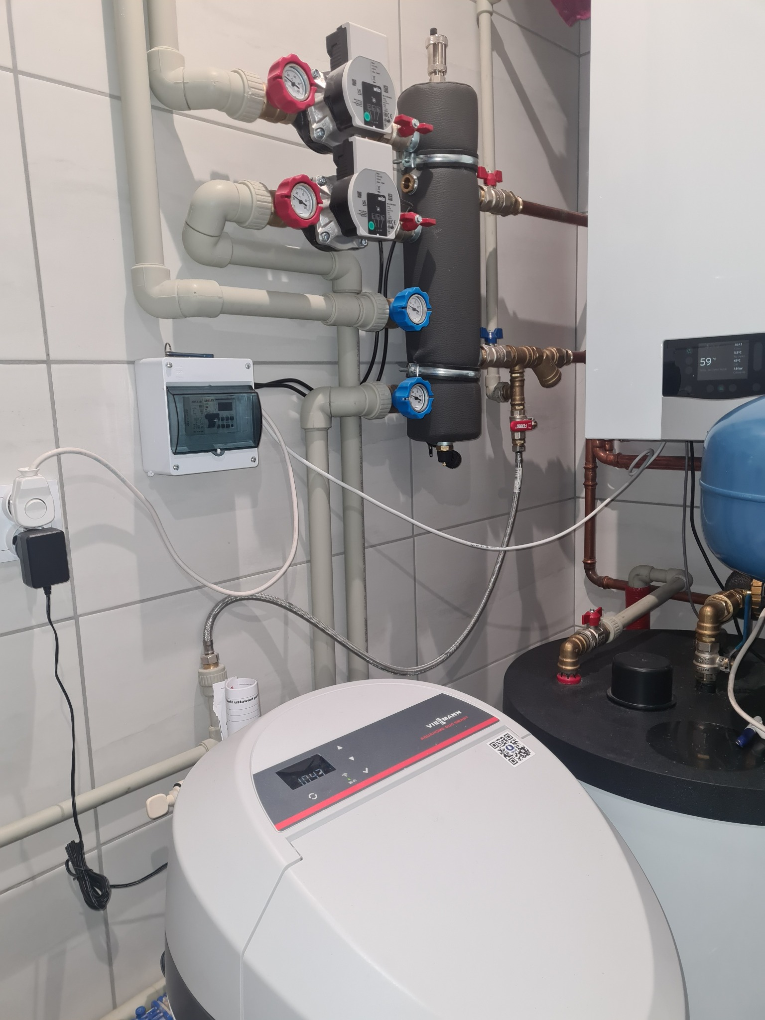 Węzeł cieplny z białym zmiękczaczem wody Viessmann Aquahome Duo Smart, widoczne rury, zawory, manometry, sterownik i zbiornik akumulacyjny, na ścianie białe płytki.