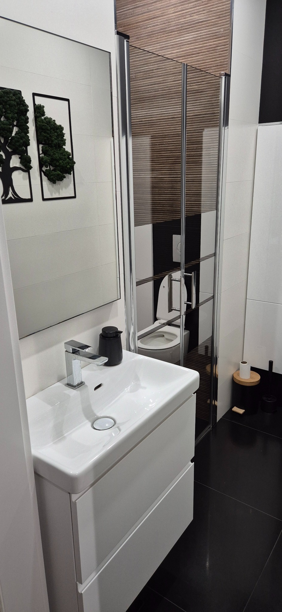 Nowoczesna łazienka z białą umywalką na szafce, lustrem i kabiną prysznicową. Dekoracje ścienne z motywem drzew. Czarno-biała kolorystyka, minimalistyczny design.