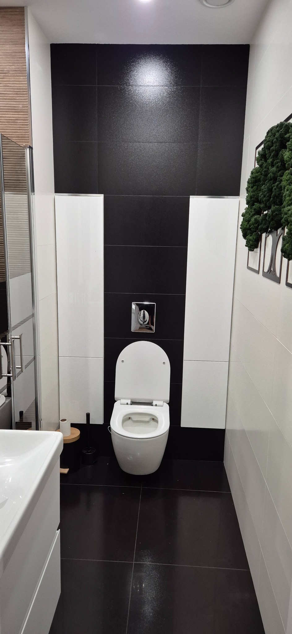 Nowoczesna łazienka w czerni i bieli z wiszącą toaletą, dekoracją z mchu i umywalką. Minimalistyczny design z akcentem natury.