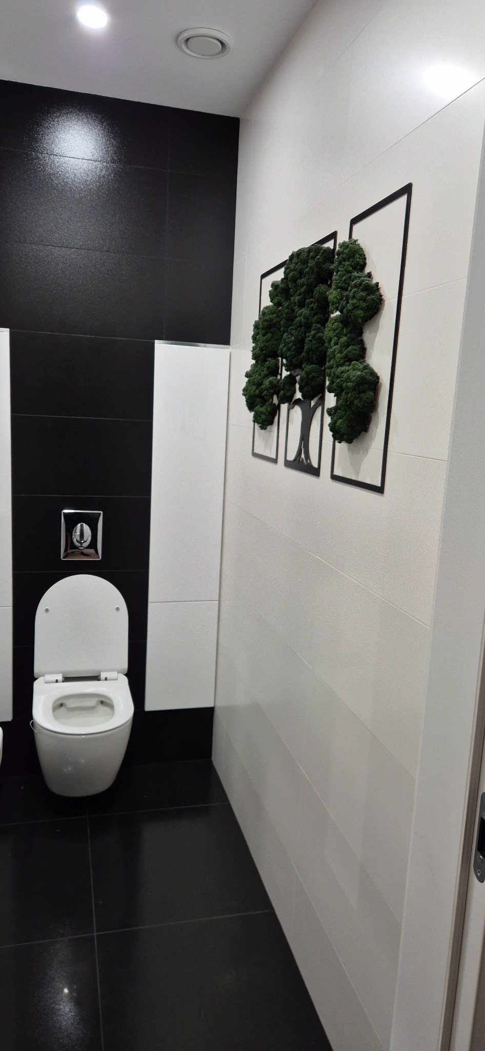 Nowoczesna łazienka z czarno-białymi płytkami i dekoracją ścienną z mchu w kształcie drzewa. Wisząca toaleta i minimalistyczna zabudowa.