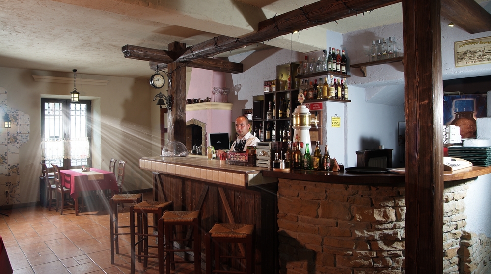 Wnętrze restauracji hotelowej z barem rustykalnym, drewnianymi belkami stropowymi i kelnerem za ladą, światło słoneczne wpadające przez okno.