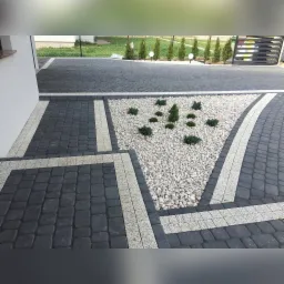 Ułożona kostka brukowa w odcieniach szarości z dekoracyjnym trójkątnym klombem wypełnionym białymi kamieniami i niskimi roślinami iglastymi.