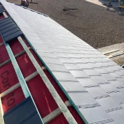 Montaż szarej blachodachówki na konstrukcji drewnianej z czerwoną membraną, widok z góry, w tle traktor i przygotowany teren pod bruk.