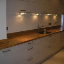 Jasnoszara zabudowa kuchenna z drewnianym blatem, nowoczesne oświetlenie podszafkowe i stalowa bateria.