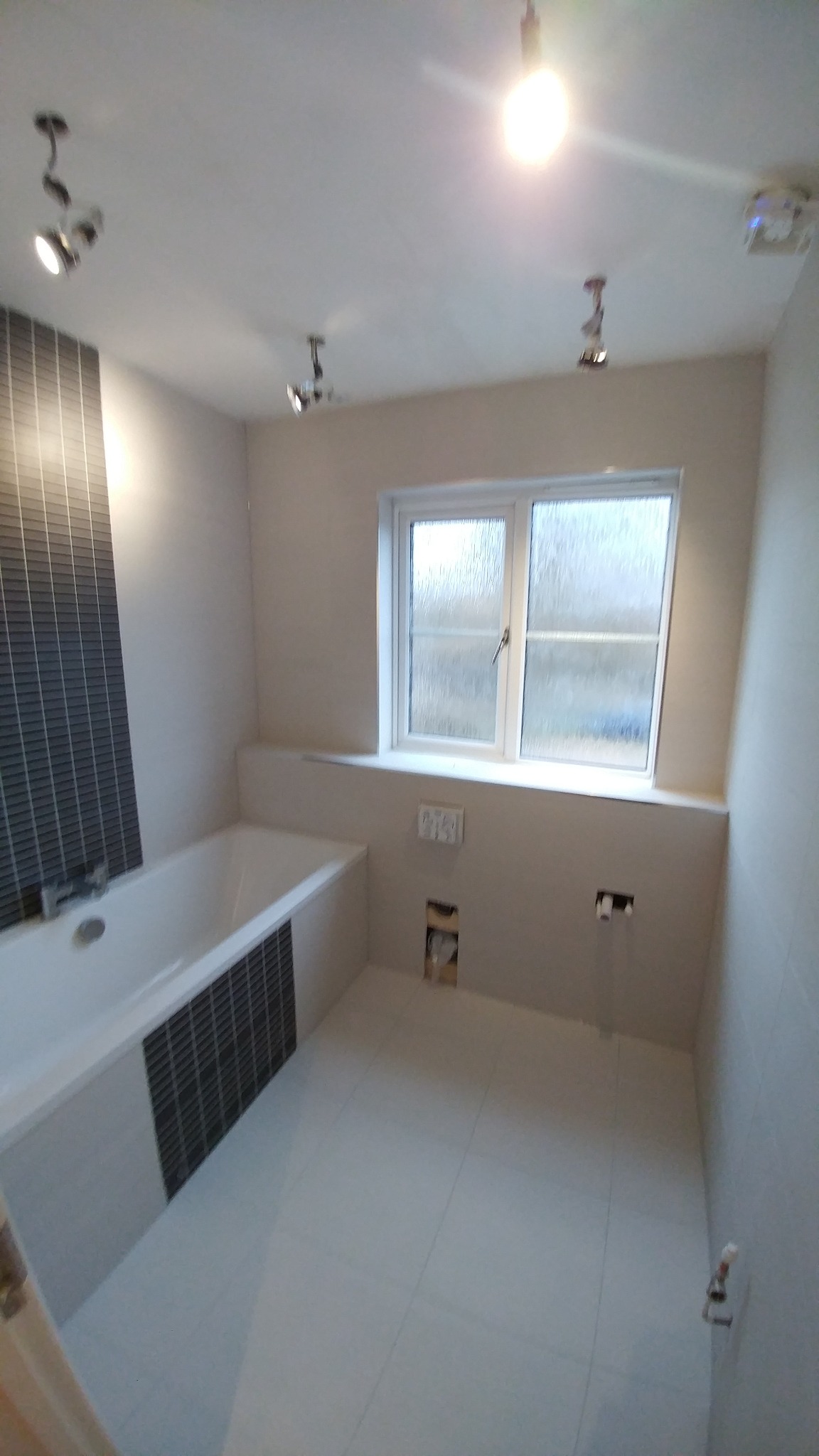 Wykończona łazienka z białą armaturą, szarymi płytkami mozaikowymi przy wannie, jasnymi płytkami na ścianach i podłodze, oraz oknem z szybą ornamentową, widoczne niedokończone przyłącza elektryczne...