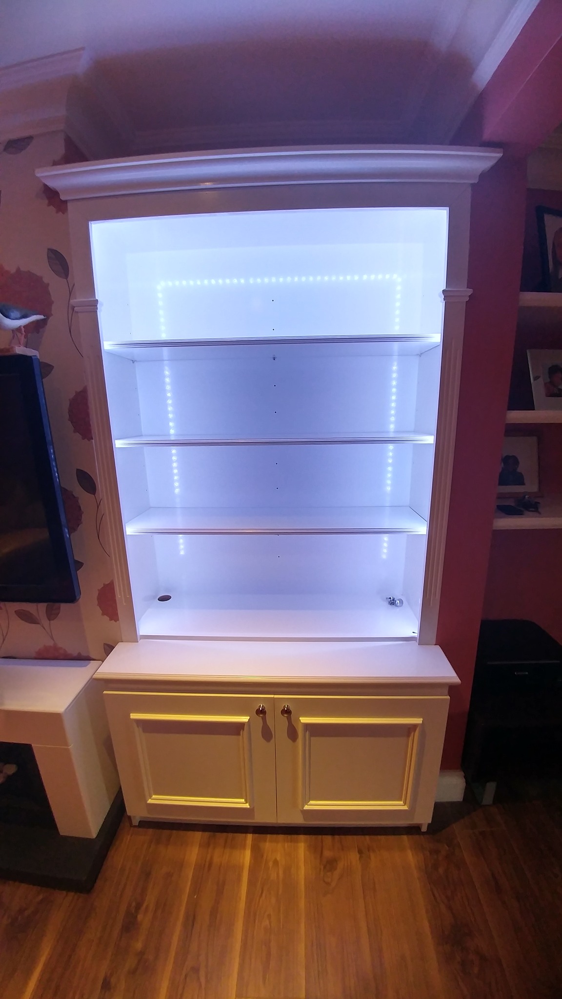 Biała zabudowa meblowa z półkami i szafką, oświetlona diodami LED, widoczna na tle różowej ściany i drewnianej podłogi.