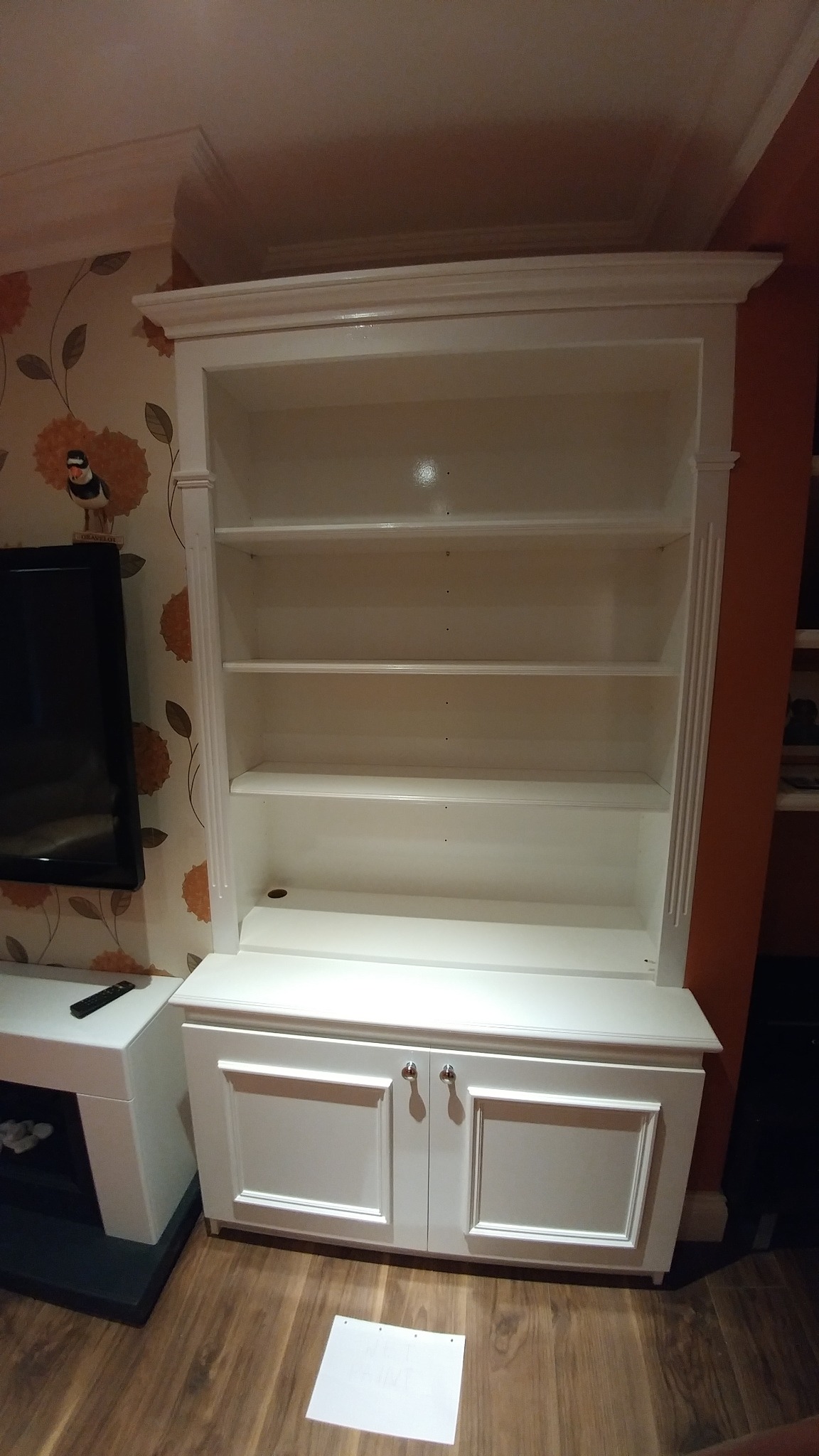 Biały regał z półkami i szafką dolną, stojący na drewnianej podłodze, z kartką papieru na podłodze, obok telewizora na białej szafce RTV, na ścianie tapeta w kwiaty.