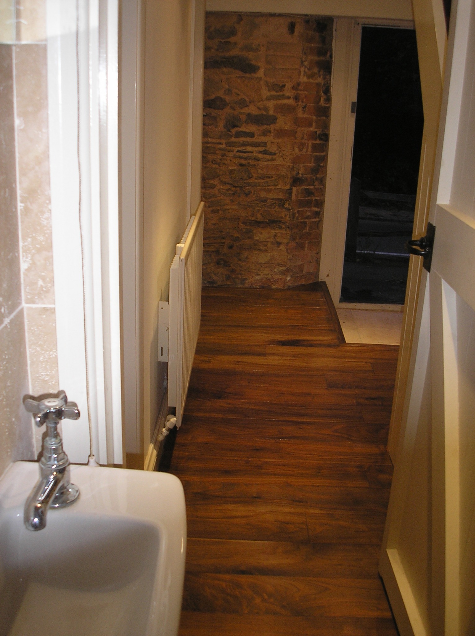 Wnętrze łazienki z drewnianą podłogą, widoczna umywalka z chromowaną baterią, grzejnik ścienny i fragment ściany z cegły za otwartymi drzwiami.